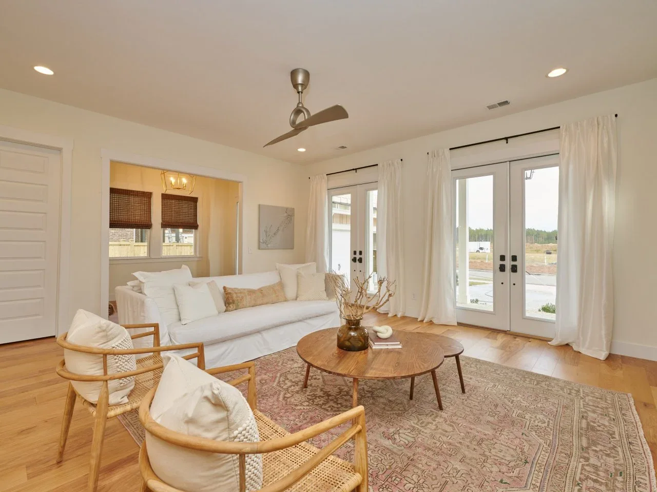A white sofa and two timber-framed woven chairs surround round timber tables beside long white curtains.