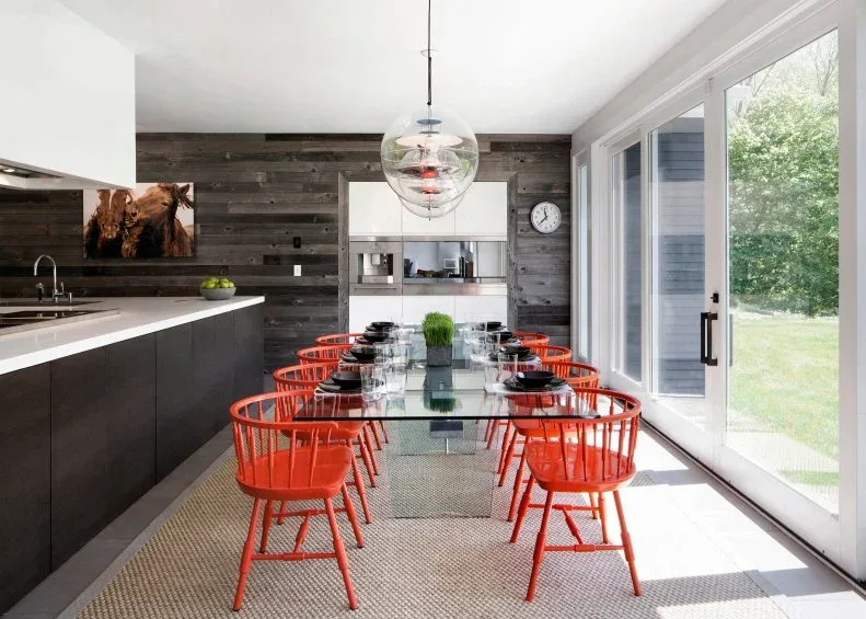 Clear rectangular dining table with orange spindle-back chairs between dark kitchen cabinetry and sliding doors