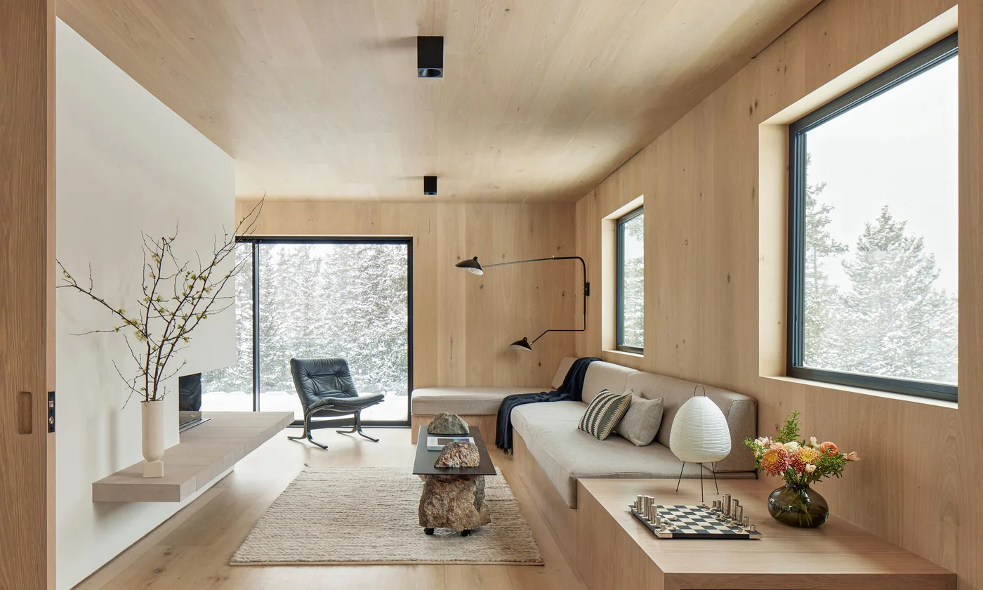 A pale wood-lined living room has a plain white fireplace wall and long hearth opposite built-in cushioned seating, with dark window frames and a textured rug.