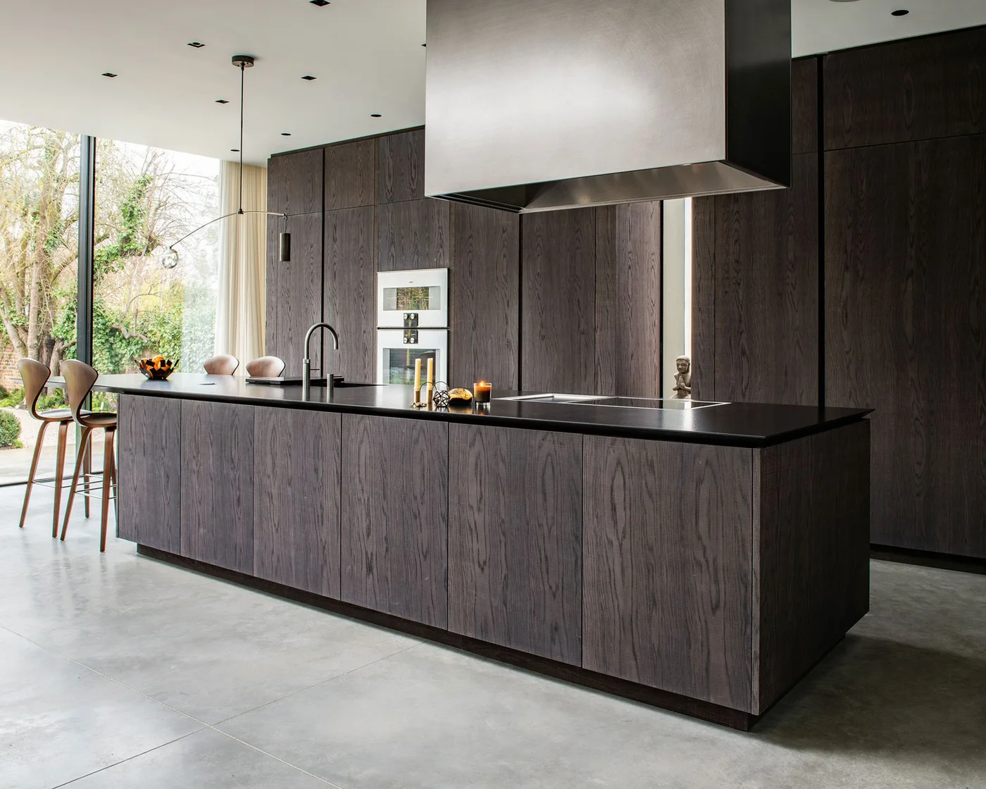 Dark rough-sawn oak cabinets and a long black-topped island sit beneath a broad metal hood beside full-height garden windows.