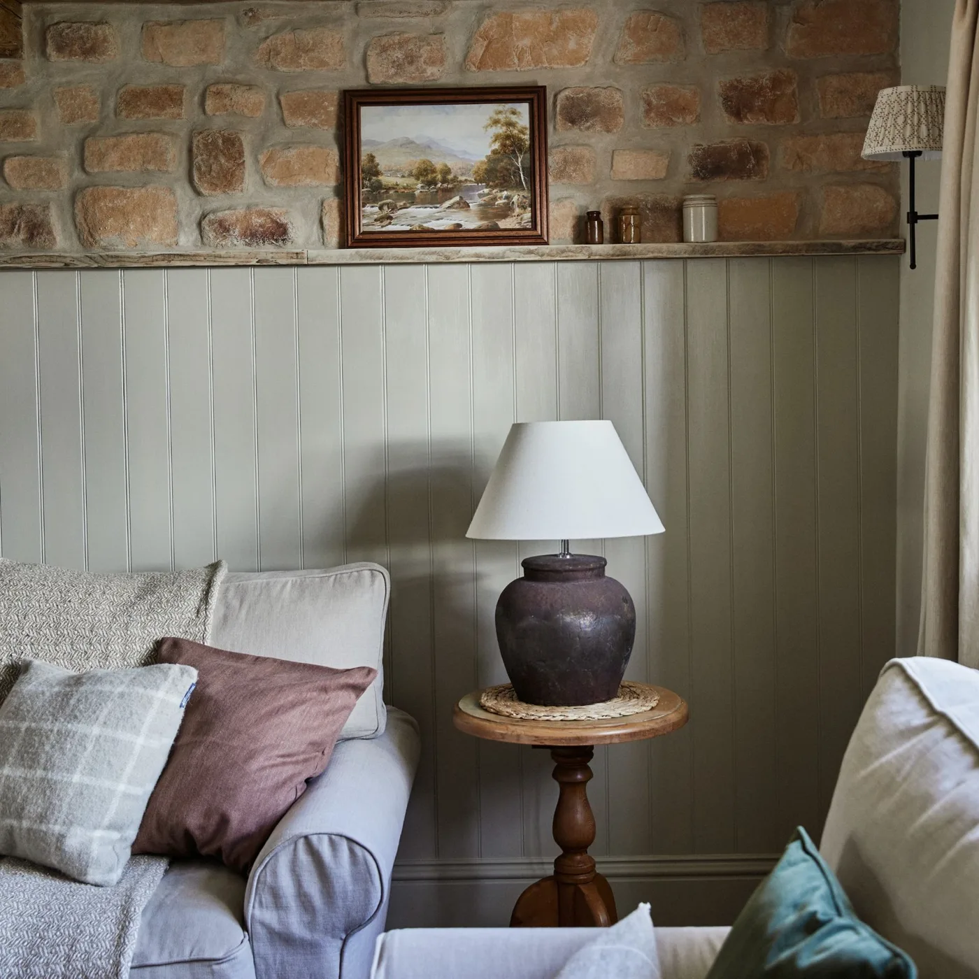 Warm irregular stone above muted lower wall panels in a living room with pale seating and a wood side table