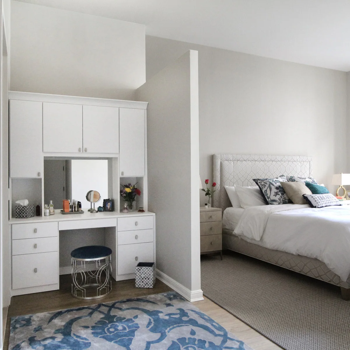 A white fitted dressing table with drawers and an overhead cupboard stands in a recess beside a pale upholstered bed.