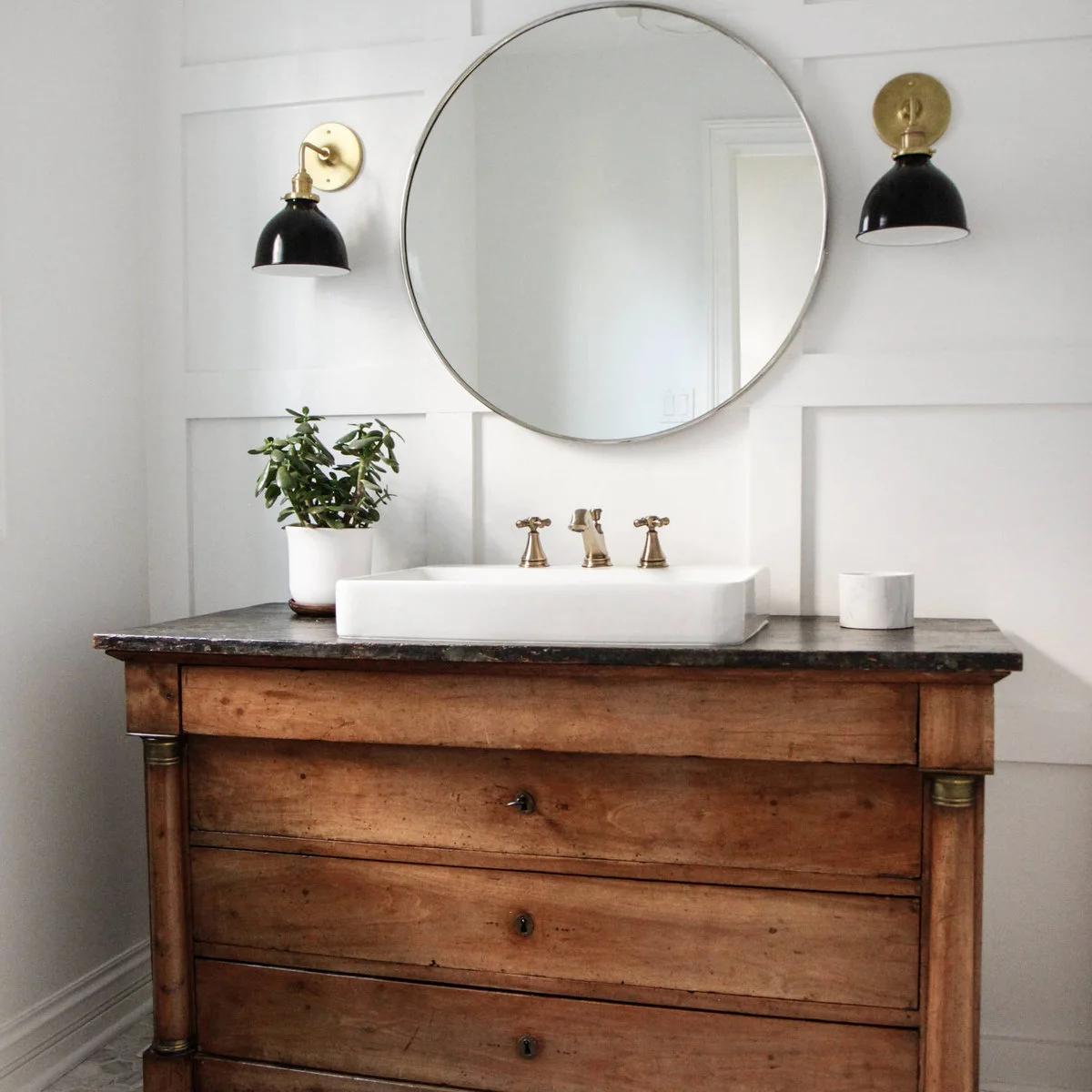 Round mirror between black wall lights above a wooden bathroom vanity