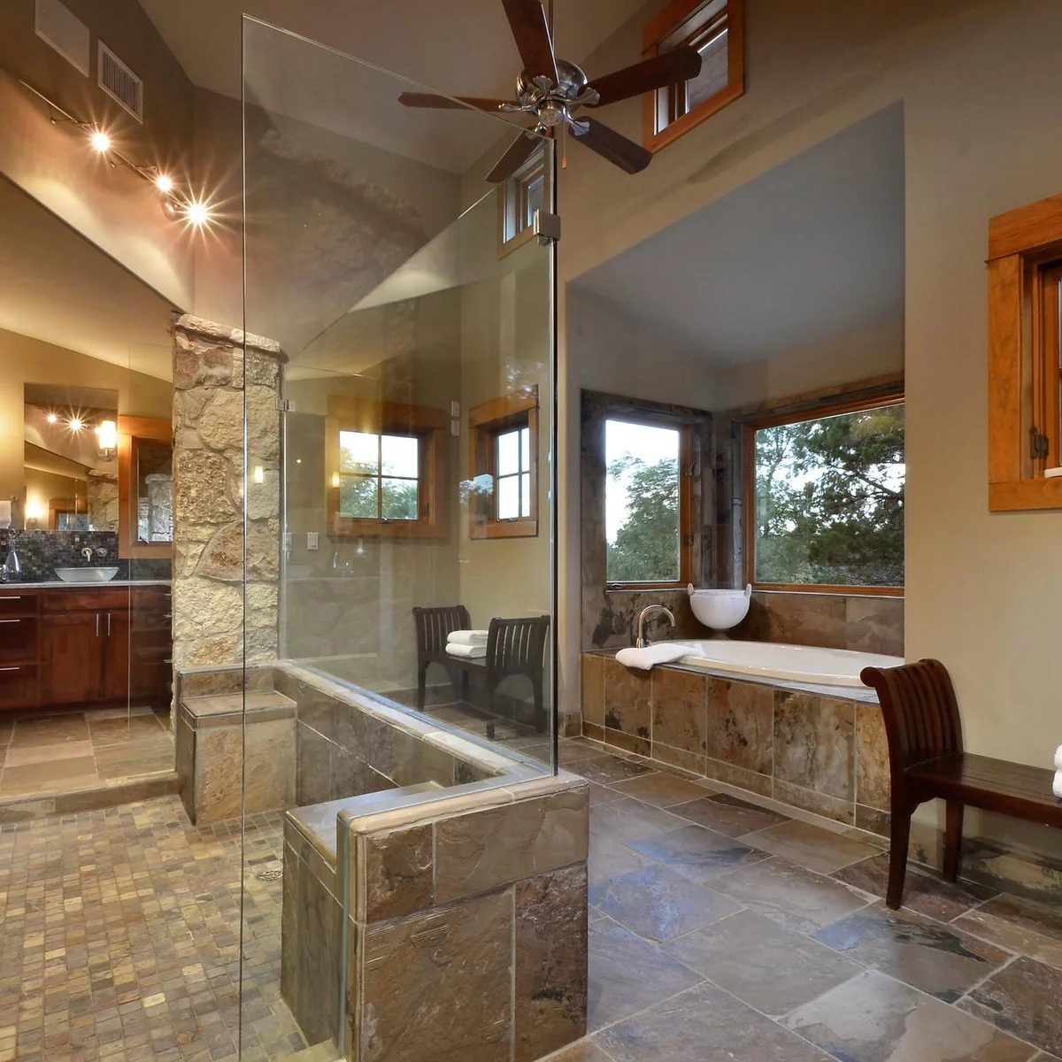 A large shower with glass panels above low tiled walls, a small-tile floor and warm brown finishes in a bathroom with high windows.