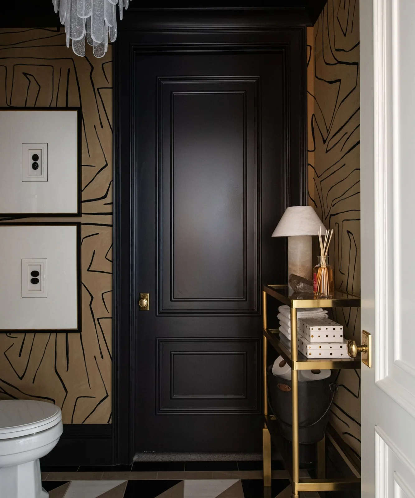Gold-colored walls with black angular lines surround a black paneled door, framed art and open console above a black-and-pale checked powder-room floor
