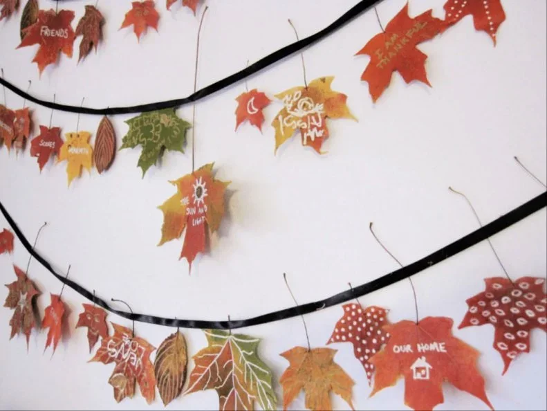 Leaf-shaped decorations carrying handwritten messages hang at varied lengths from dark cords against a pale wall