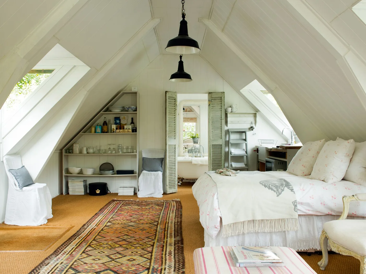 A pale attic bedroom has white roof boards, skylights, low open shelves, covered chairs, a bed and shutter-like doors to an adjoining space.