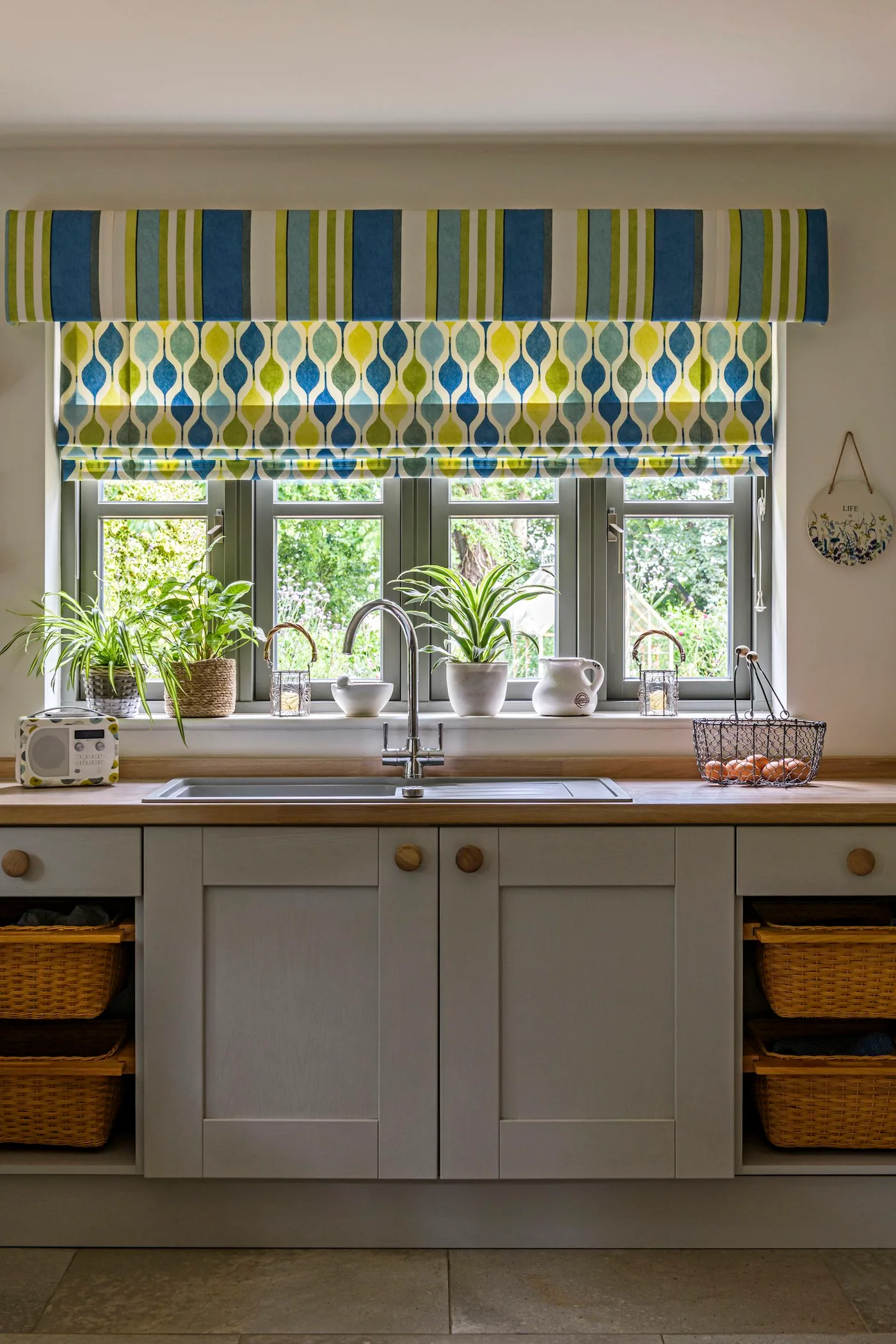 Kitchen sink beneath blue and green patterned blind, with gray cabinet fronts, wooden counter and open basket shelves.