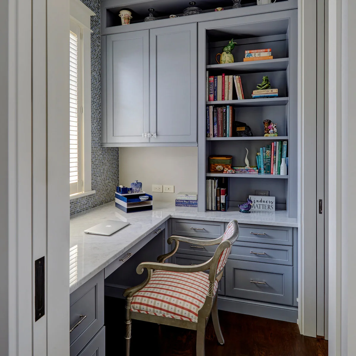 Small gray office with wraparound pale desk, closed upper cupboard, open bookcase and striped upholstered chair
