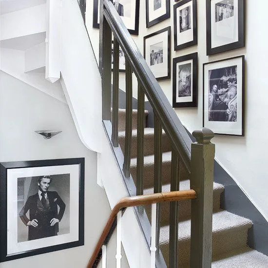 Dark picture frames with pale mounts line a white stair wall beside a dark banister, warmer handrail and pale carpeted steps