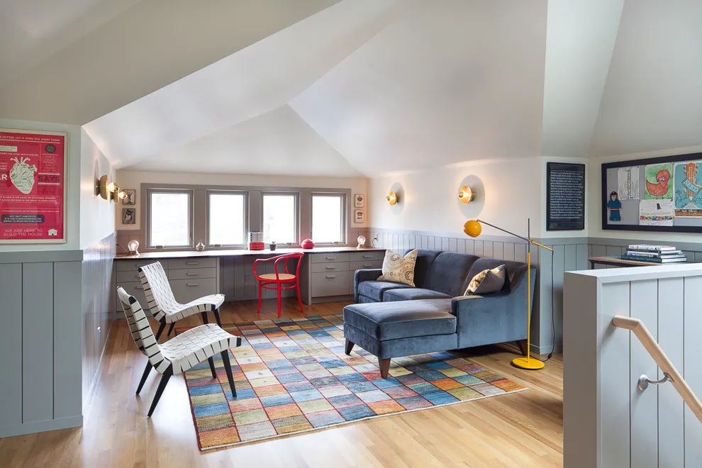 A gray fitted desk and cabinets with a red chair sit behind a blue sofa in a bright attic room