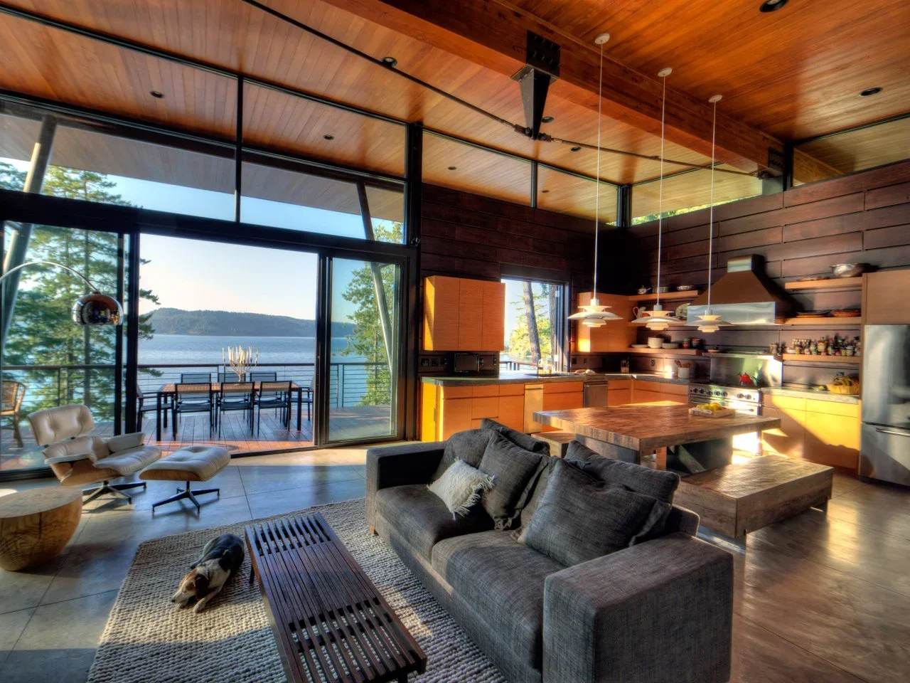 A gray sofa and dark bench coffee table sit beside a timber kitchen and dining surface facing broad water-view glazing.