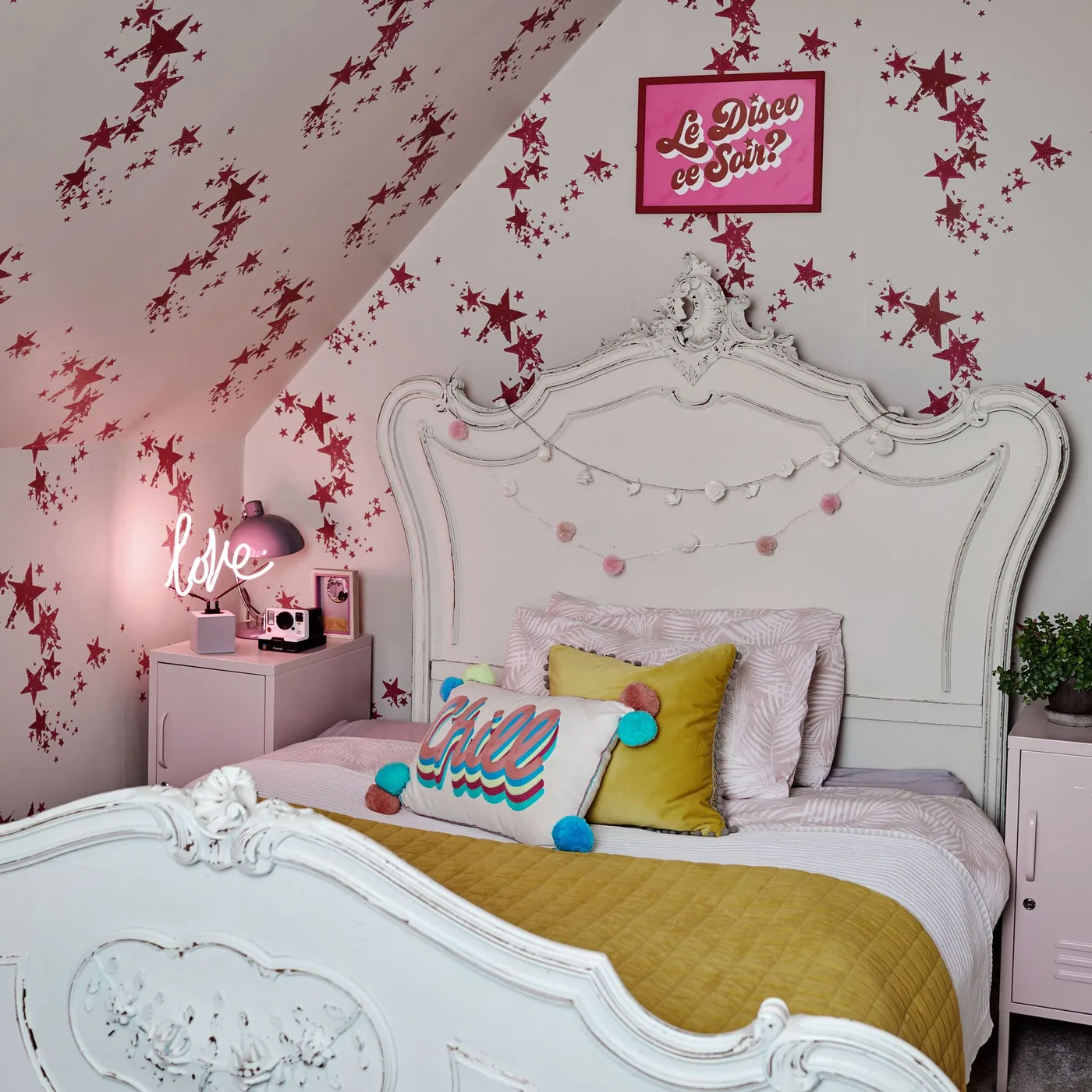Pink and burgundy stars cross a sloping bedroom wall above an ornate white bed and colorful cushions.