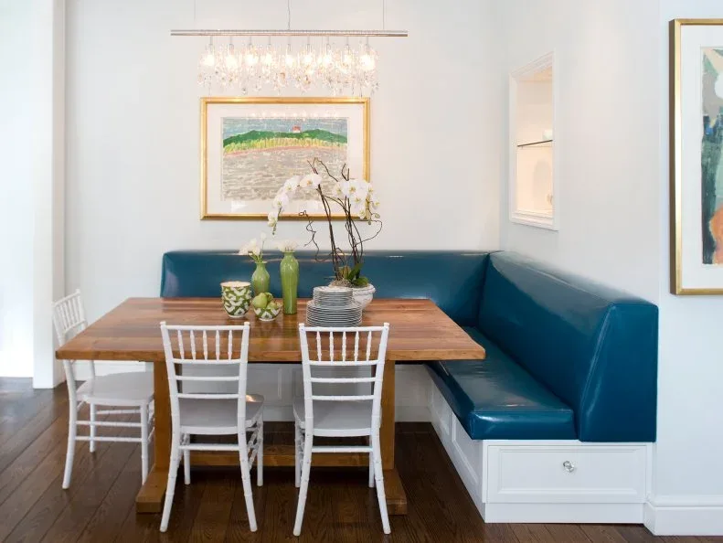 Blue fitted corner banquette with a white drawer base, wooden rectangular table, white chairs, framed art, pale walls, and a rectangular chandelier.