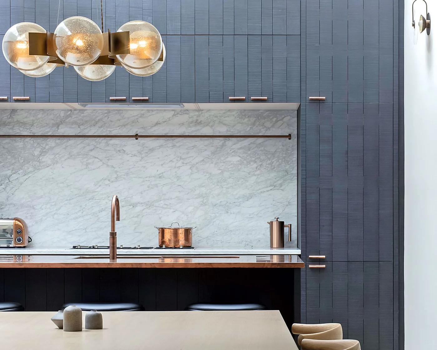 Kitchen with blue-charcoal vertically textured cabinets, pale veined backsplash, slim warm metal pulls, copper-colored island edge and tap, and globe lights.
