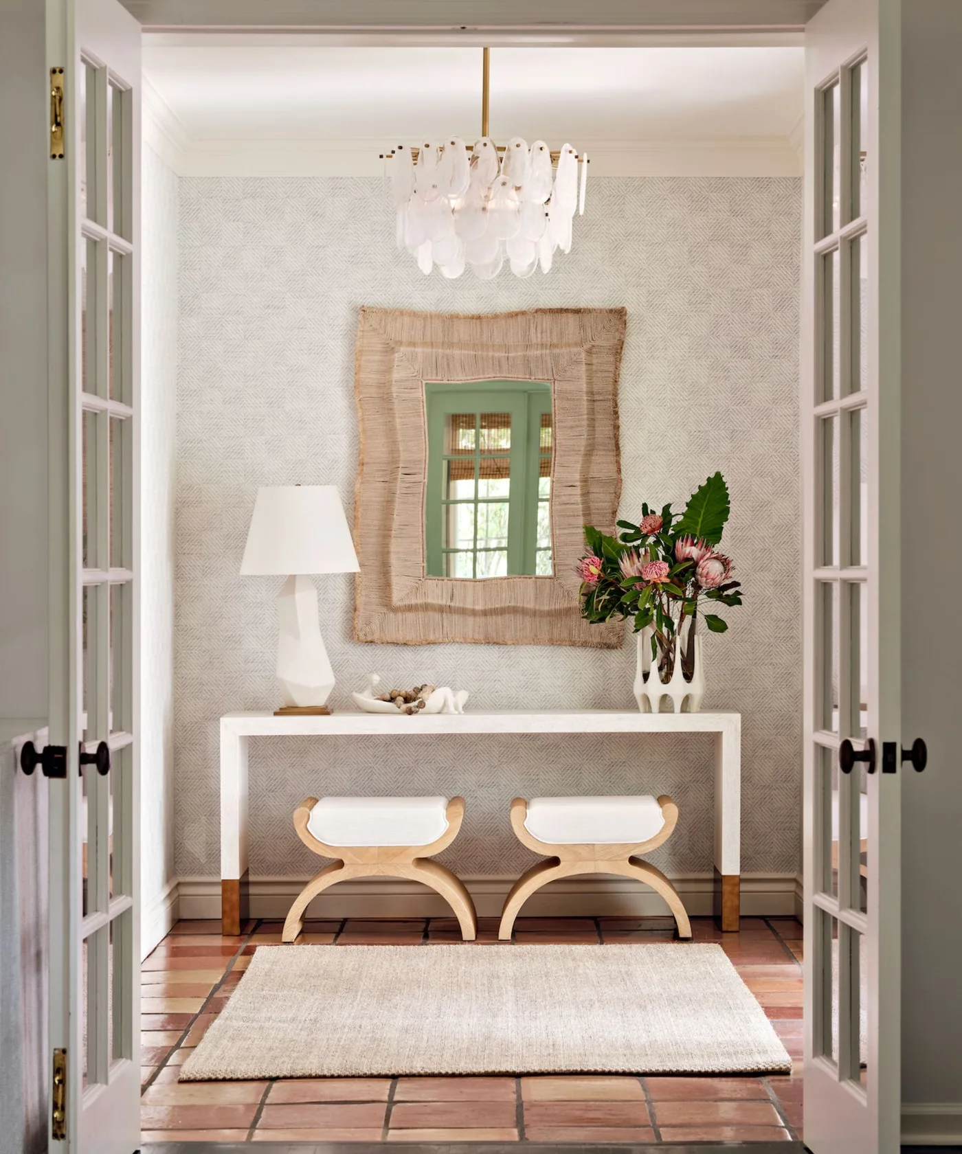 Pale entrance with white console, two curved stools underneath, large textured mirror, lamp and glazed double doors.