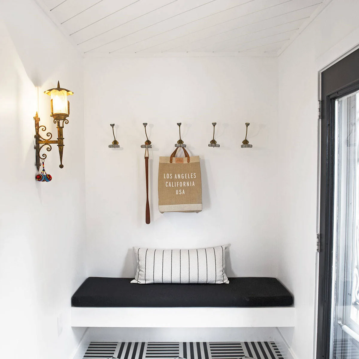 Small white entry nook with a dark bench cushion, simple coat hooks and a black and white patterned floor.