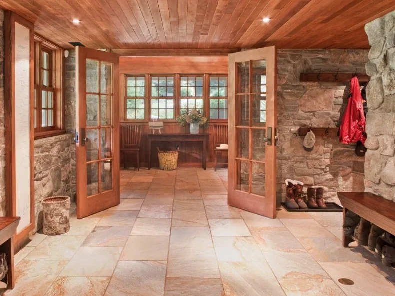 Wood benches with shoe storage line a stone-walled mudroom around open glazed doors and a broad tiled floor.