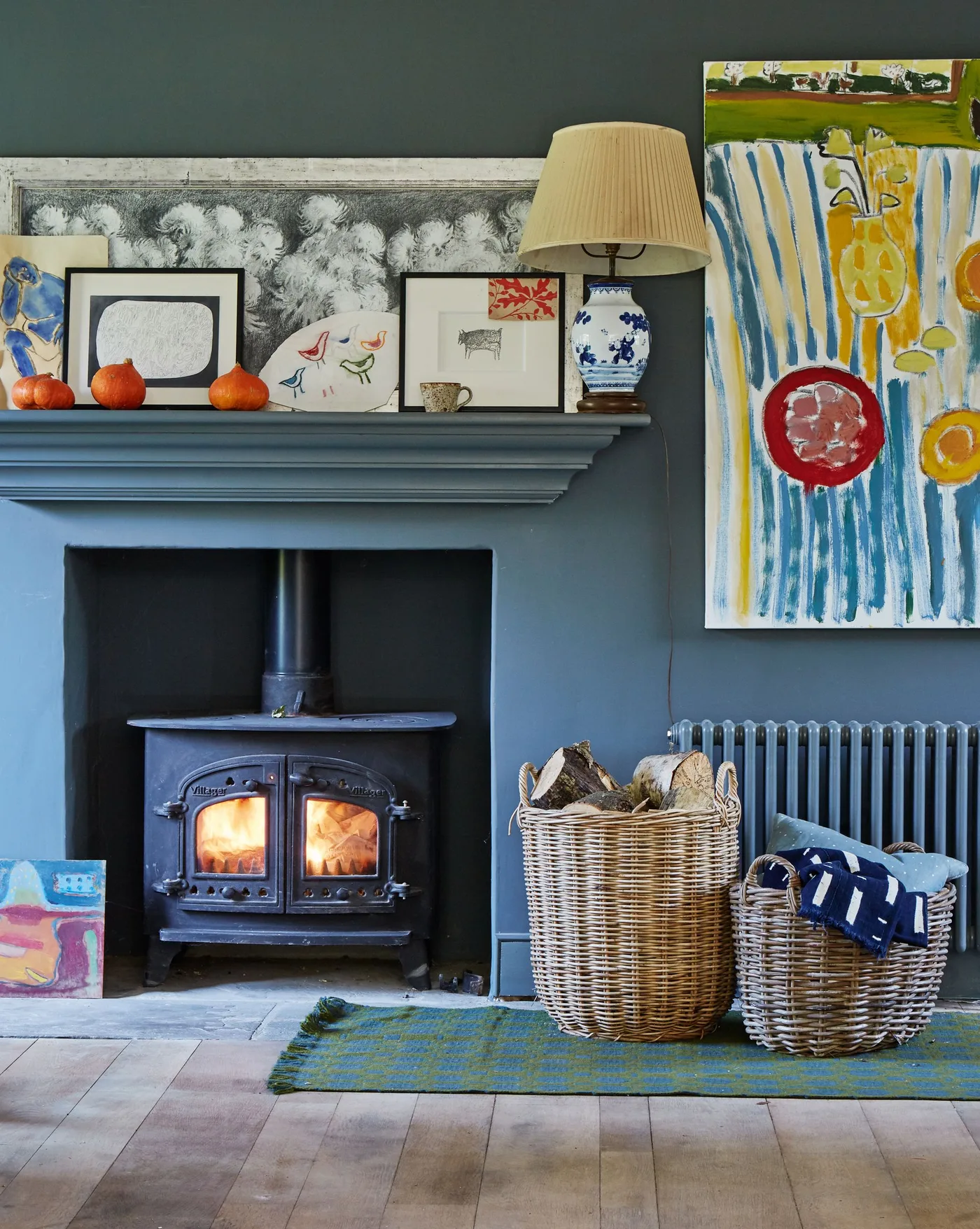 Dark stove beneath teal mantel with teal walls, pale floor, lighter rug, nearby woven baskets and stacked logs.