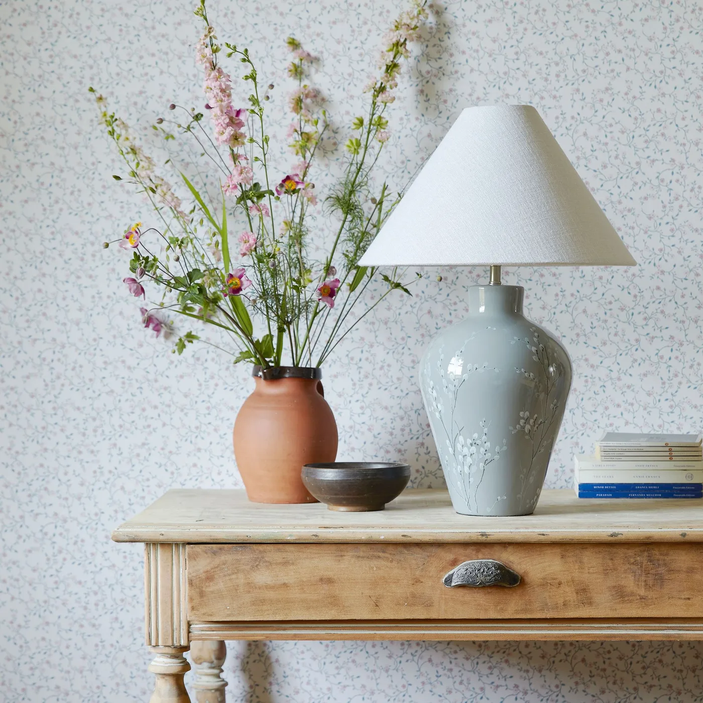 Console detail with a wooden drawer table, pale patterned wall, ceramic-based shaded lamp, vase of flowers, small bowl and books.