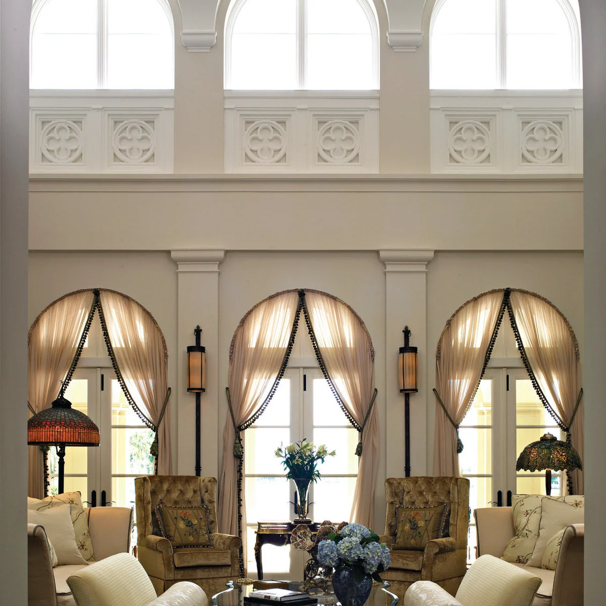A tall cream living room with arched lower doors, gathered curtains, an upper row of arched windows and traditional seating.