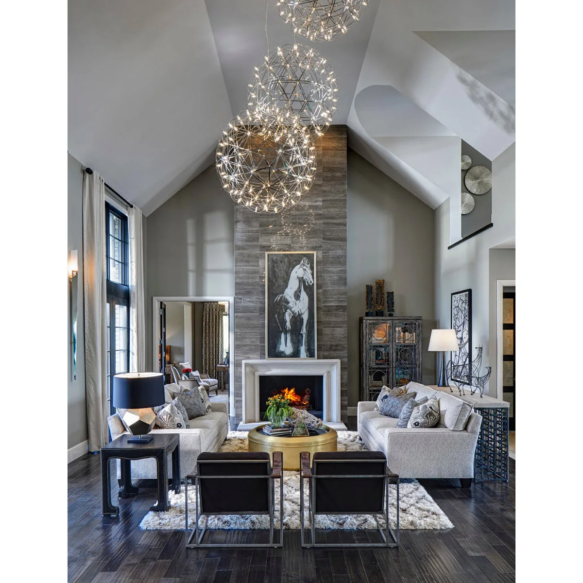 A tall gray living room with three suspended globe lights, facing pale sofas, a gold round table and a high fireplace wall.