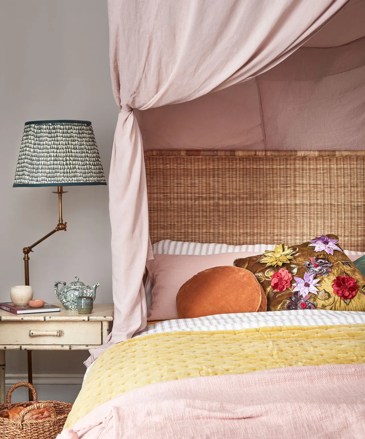 A woven headboard sits beneath pale pink canopy fabric beside a patterned lampshade.