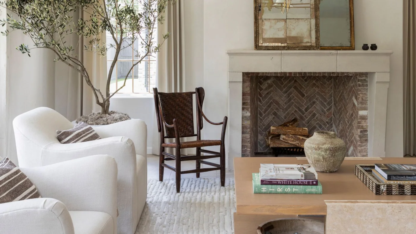A pale rectangular mantel frames a dark herringbone brick fireplace beside white curved chairs, a dark wood chair and one rectangular mirror
