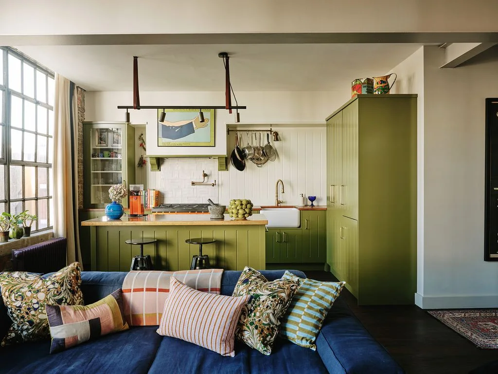 An olive-green kitchen with a short island sits immediately behind a dark-blue sofa and patterned cushions.