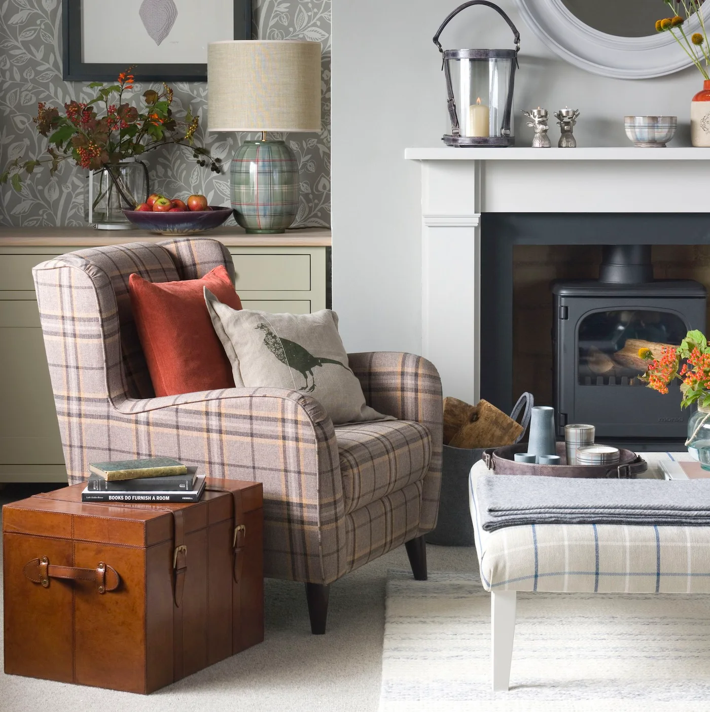 A pale plaid chair sits beside a wooden chest, a dark stove and a pale upholstered footstool