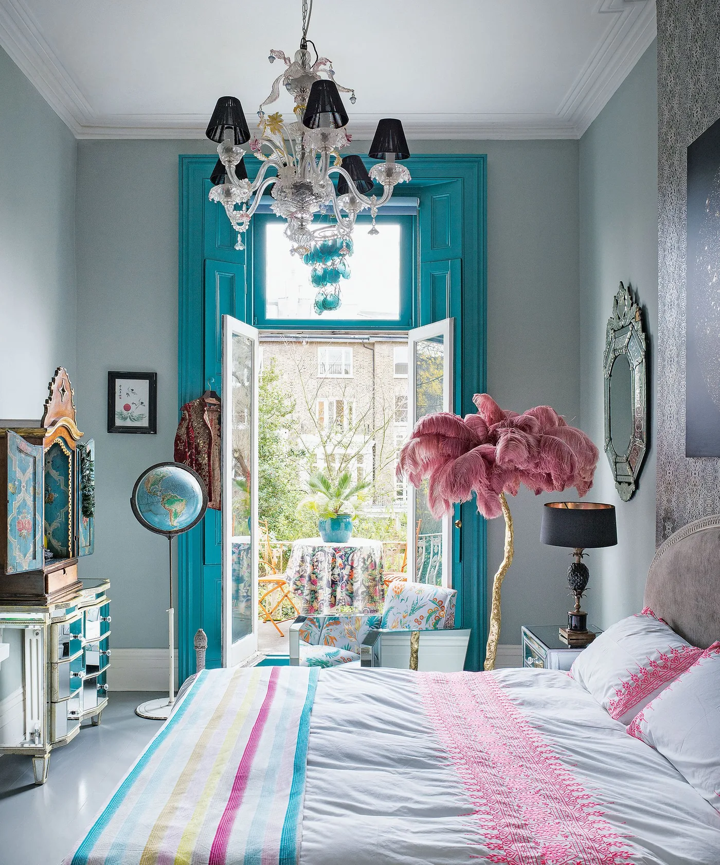 Bedroom with turquoise window trim, pale striped bedding, ornate overhead light, pink feather-shaped decoration, globe, mixed cabinets and small patterned chair.
