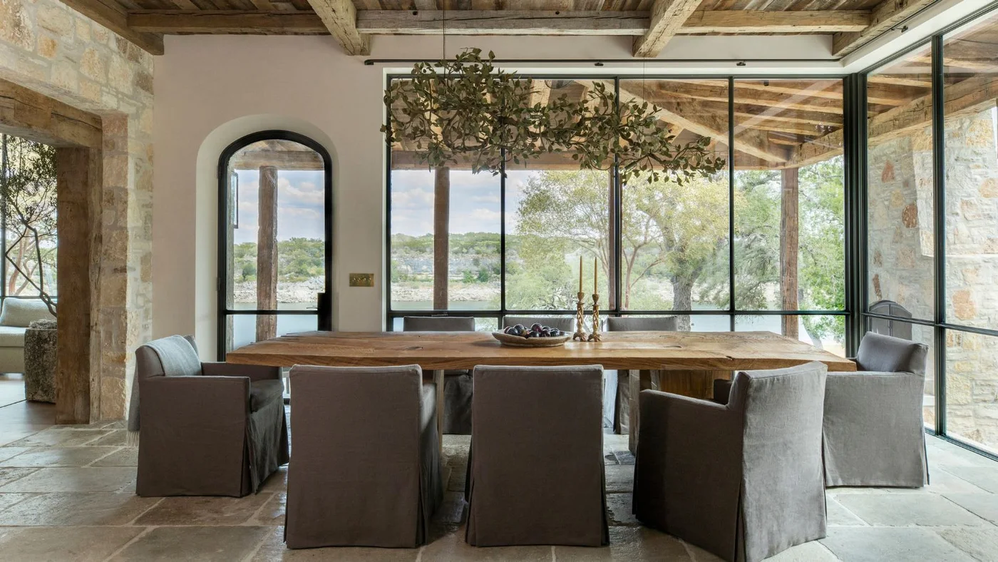 Gray skirted dining chairs surround a long wood table beneath timber beams beside broad black-framed windows.