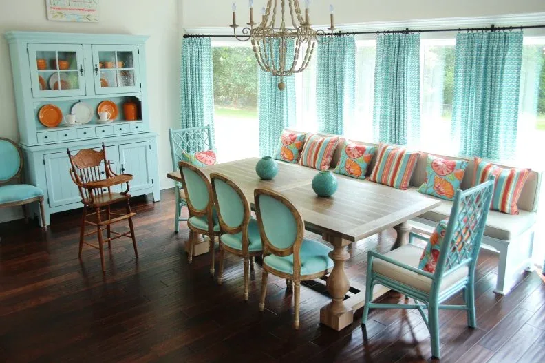 Dining room with aqua hutch and chairs, pale table, turquoise curtains and colorful cushions along a window bench.