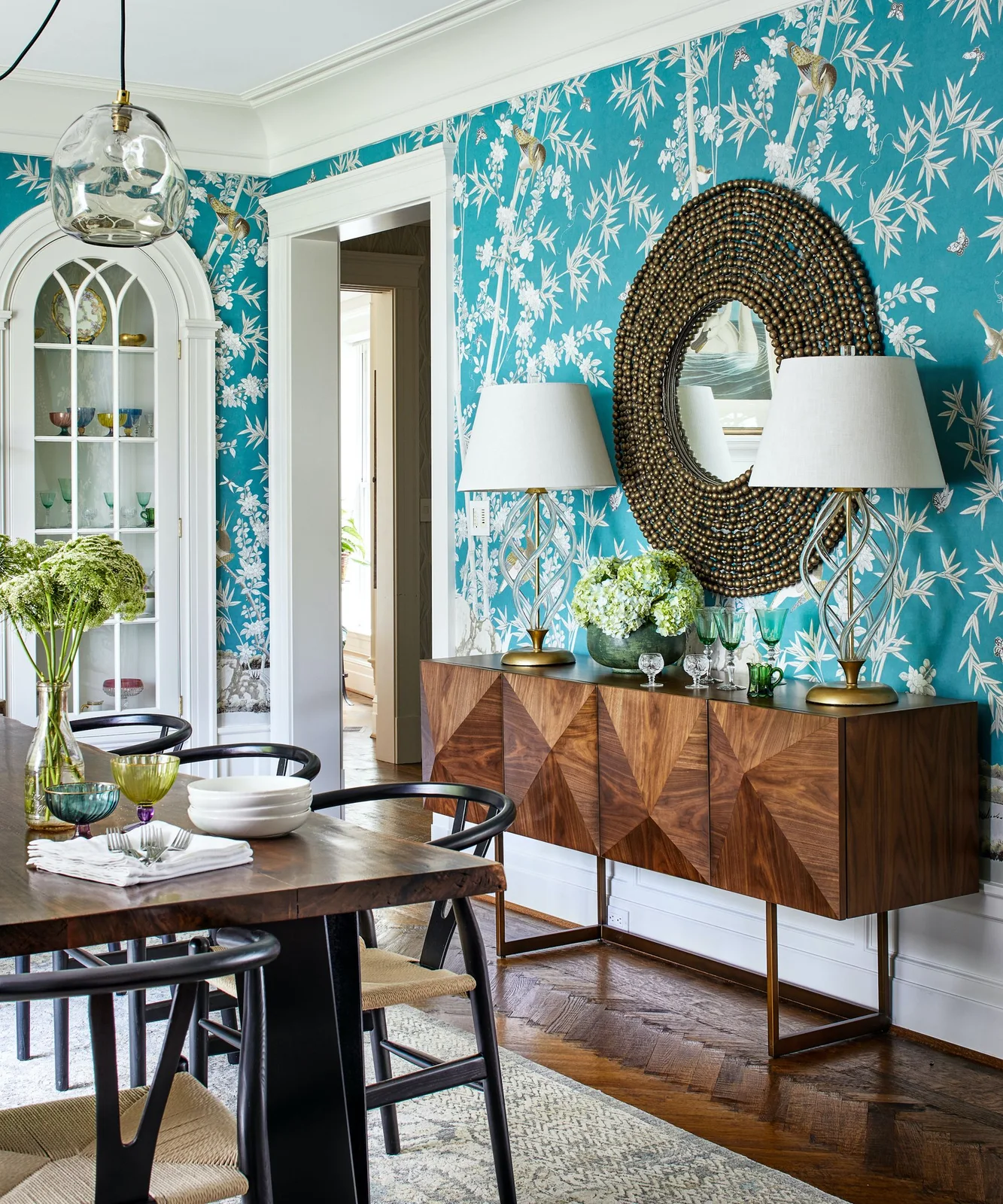 Two matching lamps stand at the ends of a geometric wood sideboard, with green and clear glasses and pale flowers between them against turquoise wallpaper.