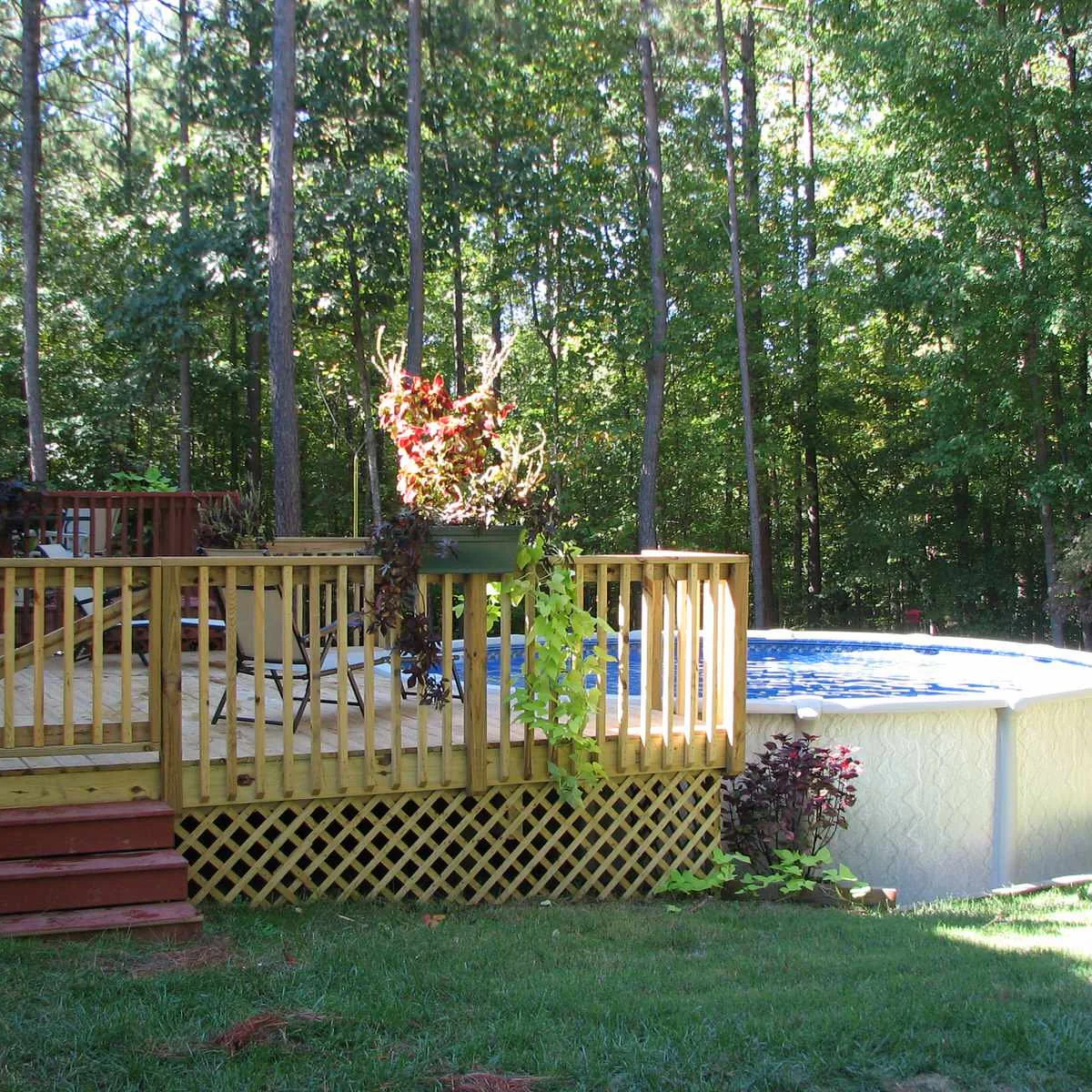 Round white above-ground pool beside a wood deck with lattice skirting, red steps and a hanging planter.