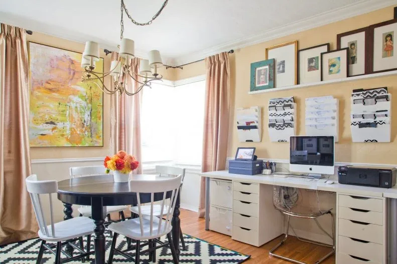 Round dining table with pale chairs beside a long white desk holding a computer and printer