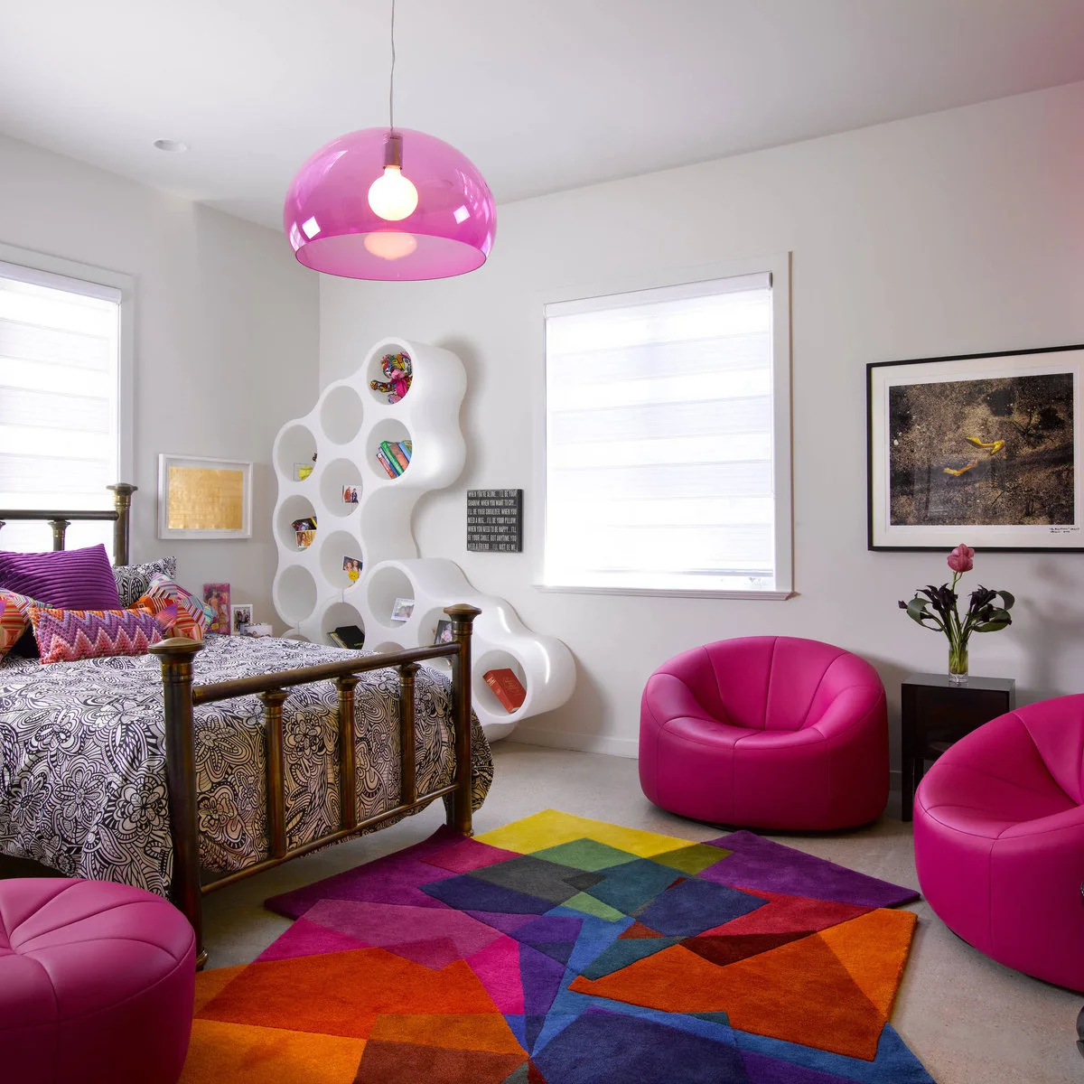 An angular multicolored rug sits beside a brass-toned bed and rounded pink chairs in a white bedroom.