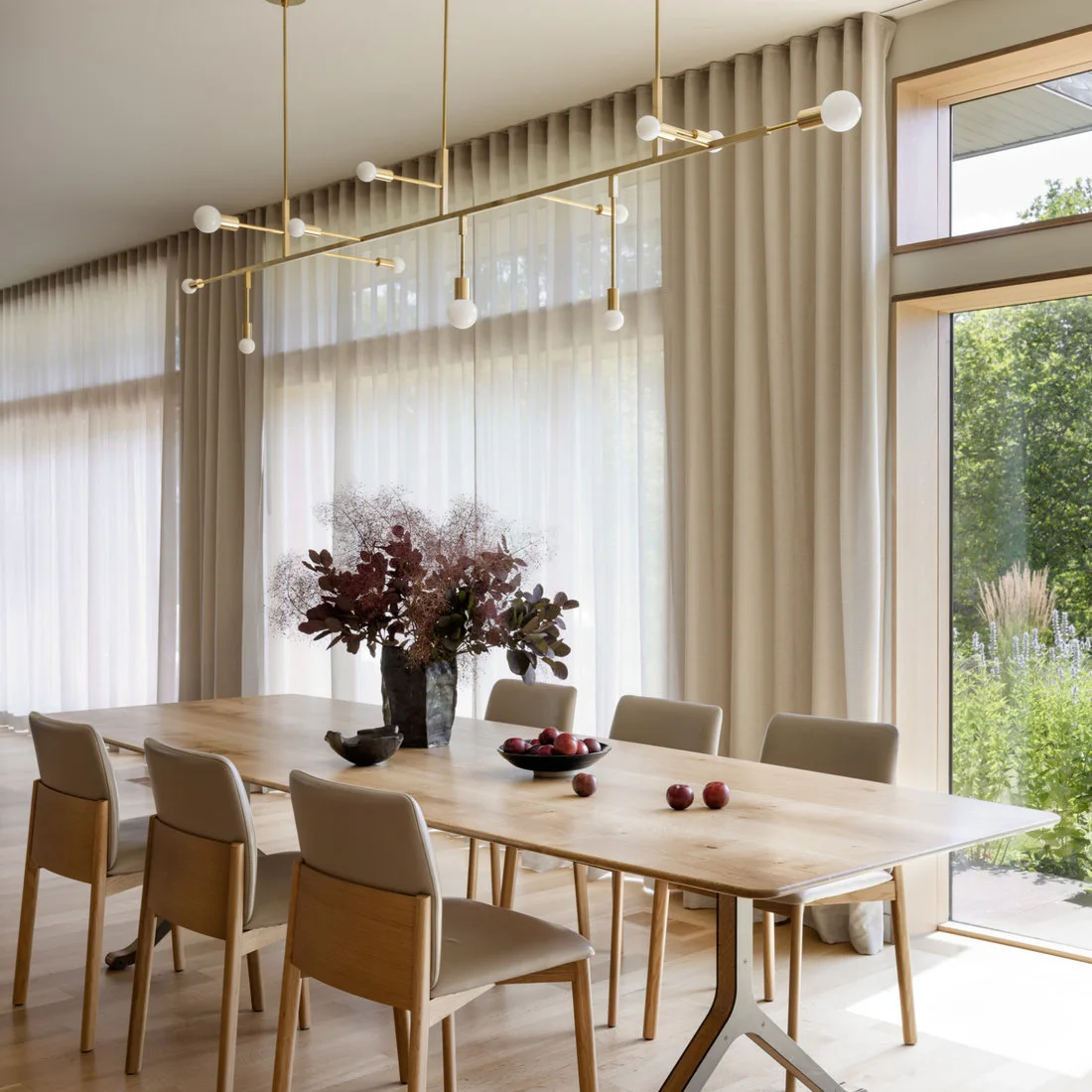 A pale rectangular dining table with angled metal supports and pale upholstered chairs stands beneath a long warm-toned bar of white globes beside tall curtains