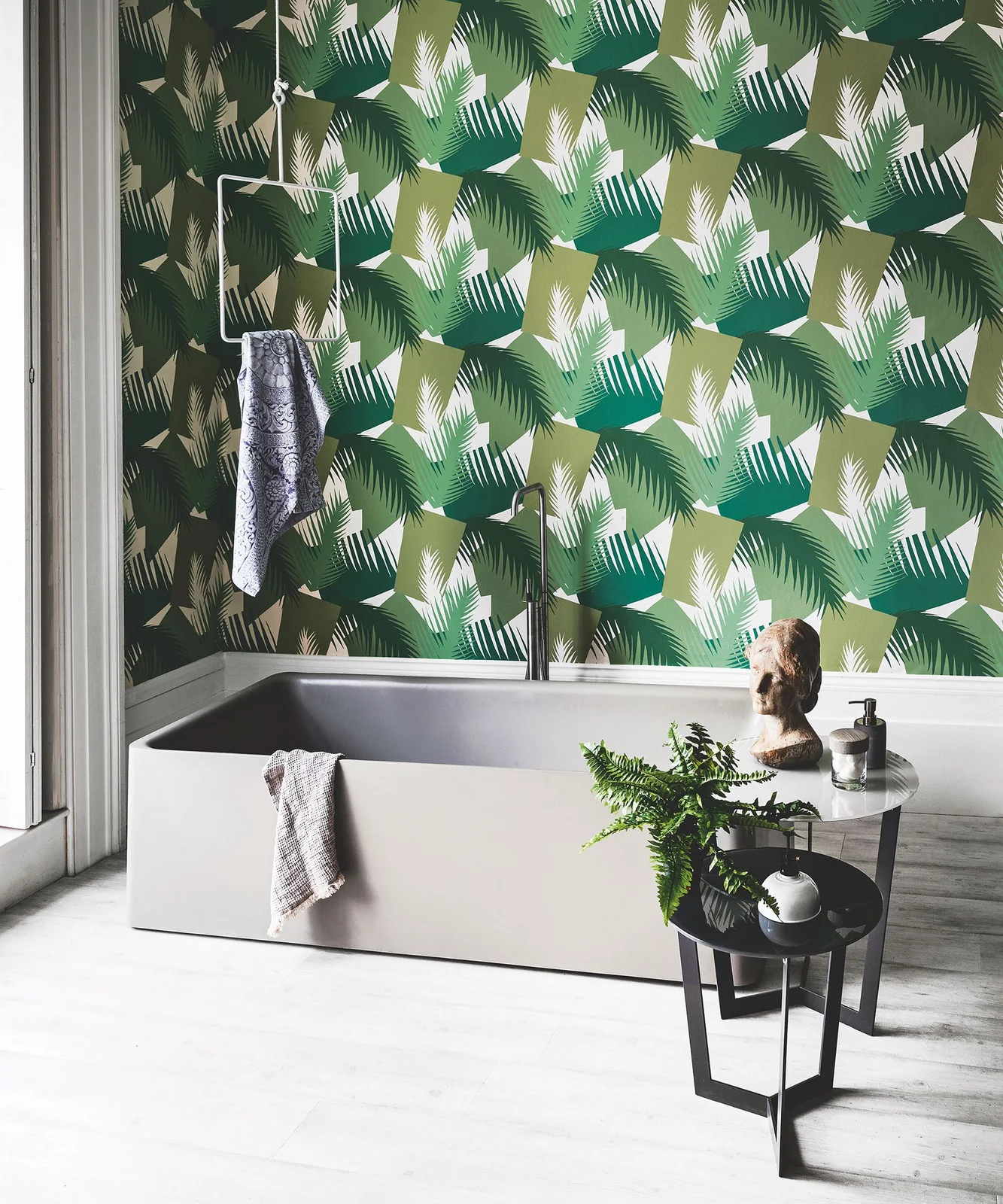 A plain rectangular bath sits below a bold green leaf-patterned wall beside dark nested tables, a plant and a hanging towel rail