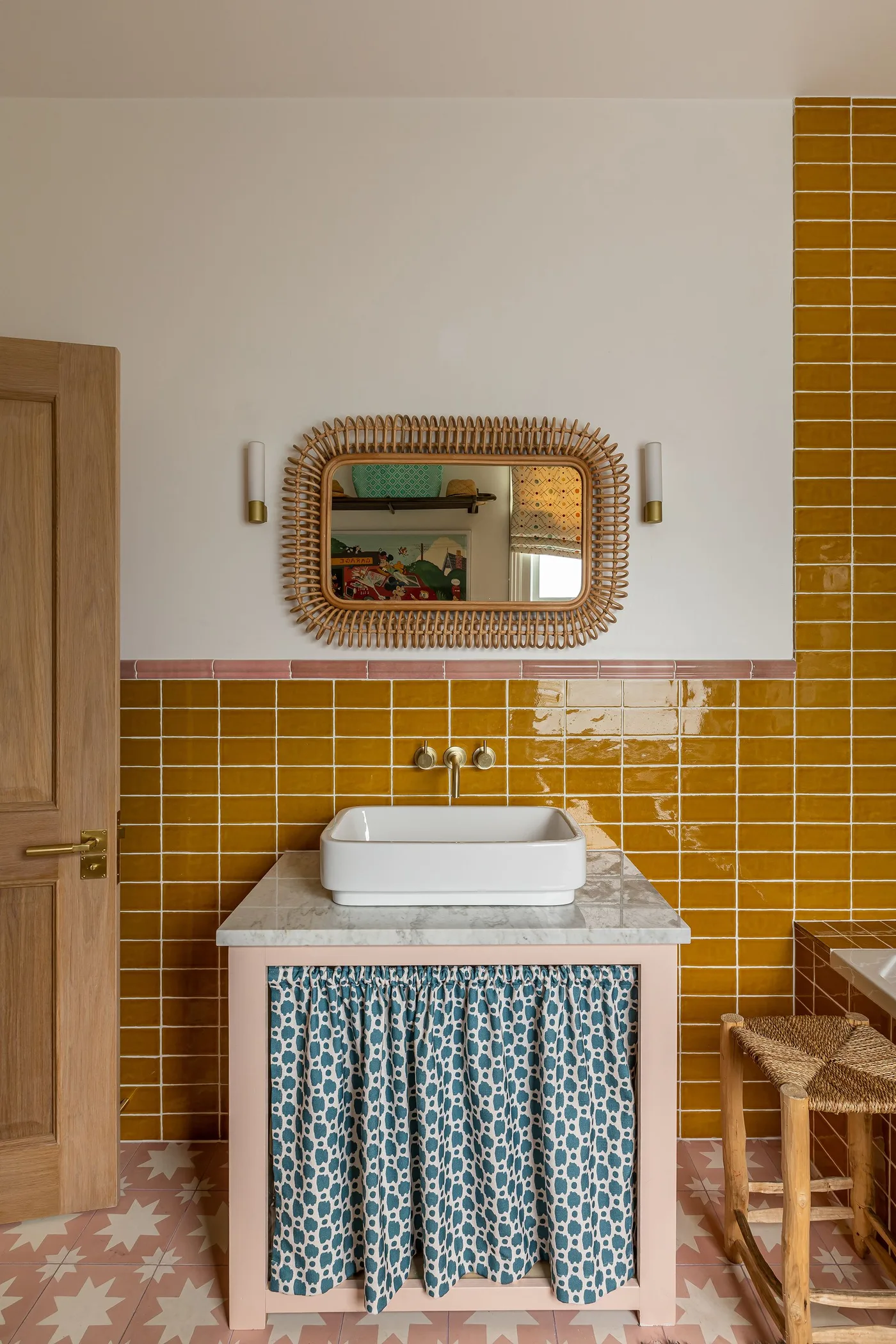Bathroom basin corner with mustard rectangular tiles, blue patterned basin skirt, woven mirror frame and pink patterned floor.
