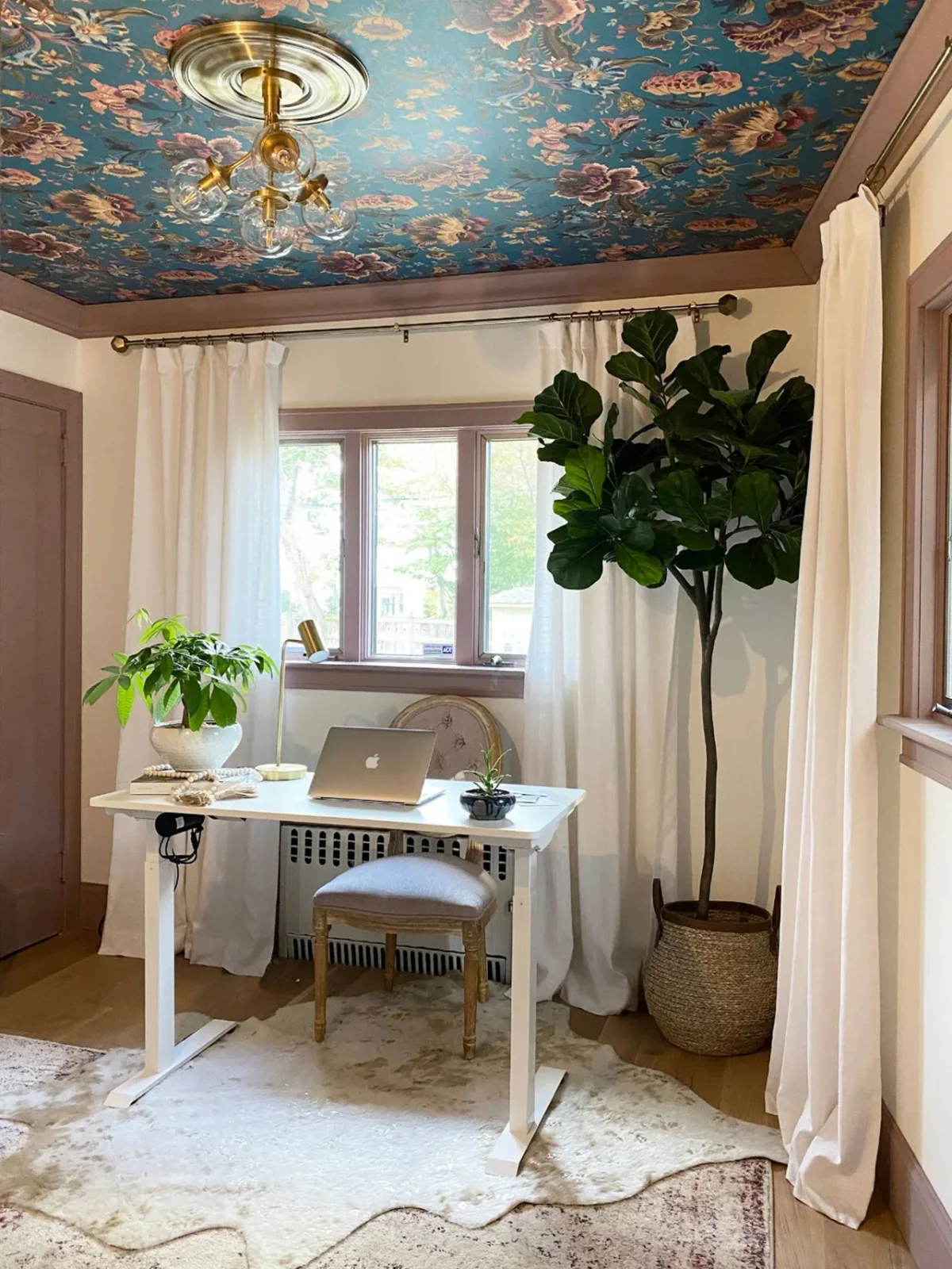 A small pale desk and upholstered chair sit under a blue floral ceiling with pink trim, pale curtains and plants beside a window.