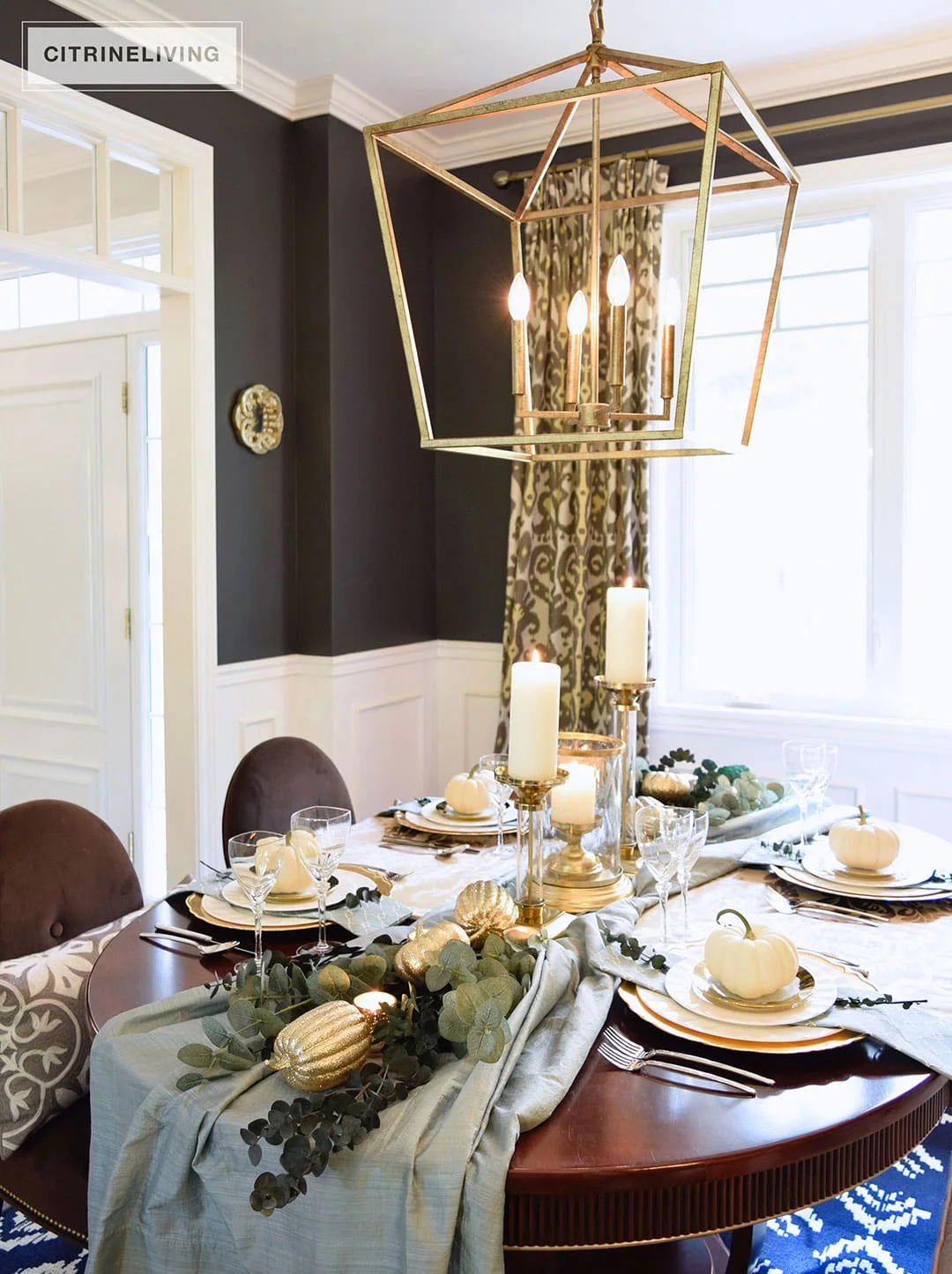 A dining table carries muted blue cloth, white and gold-colored pumpkins, greenery and tall white candles beneath a gold-colored pendant