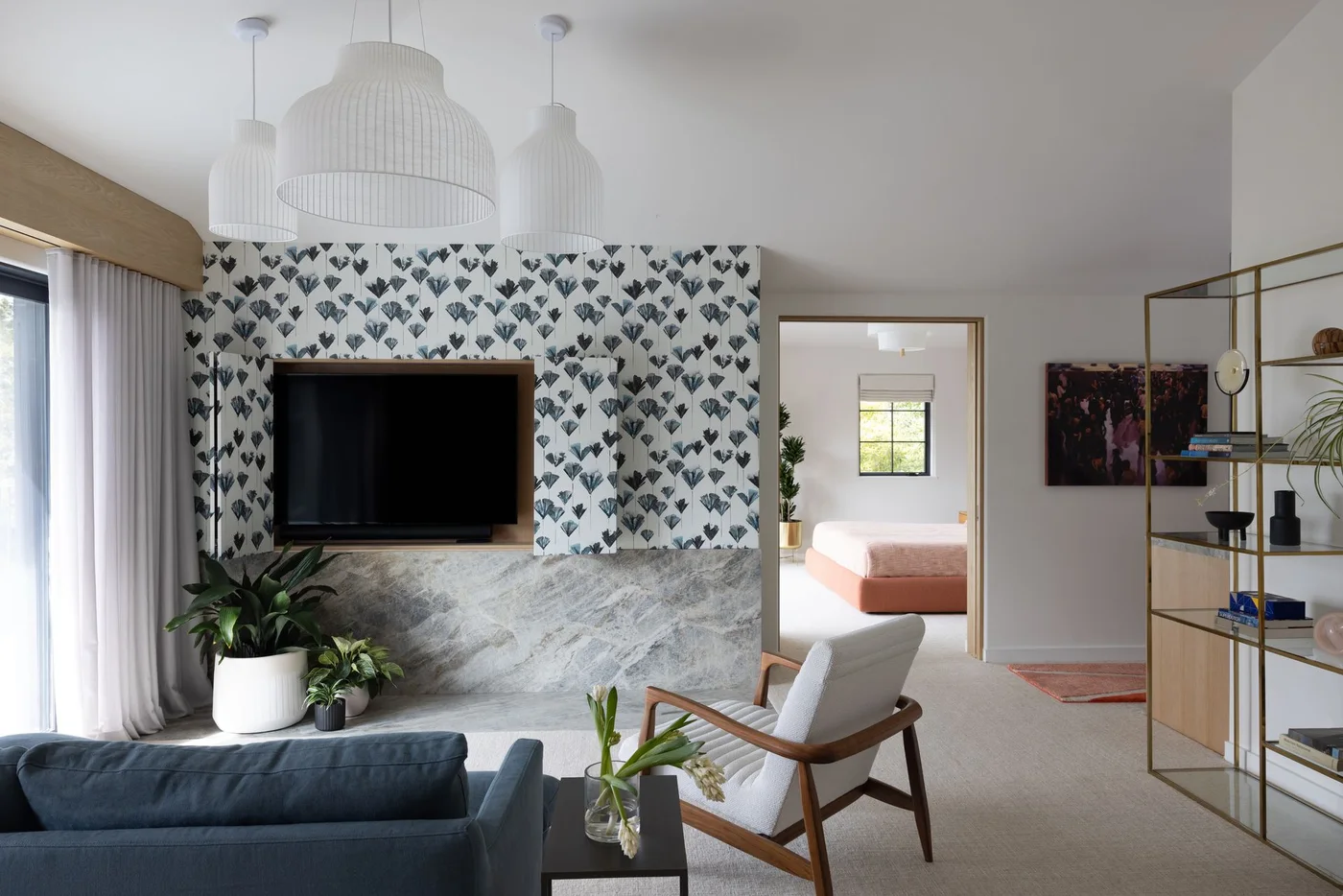 A television sits within a wood-edged recess in a botanical-patterned wall above a veined pale base beside a blue sofa