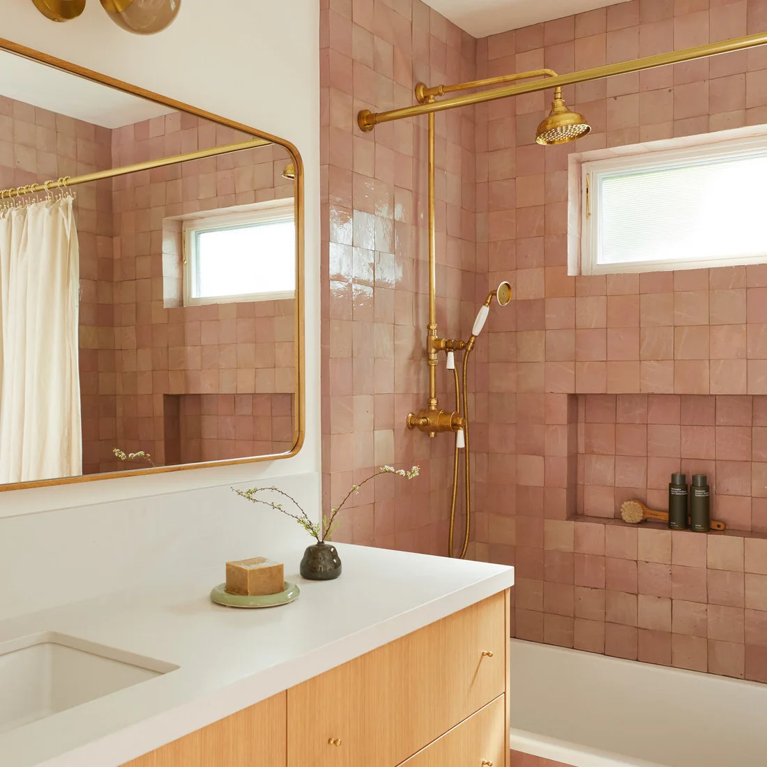 Pink square shower tiles surround a recessed niche beside a gold-colored shower fitting and pale curtain