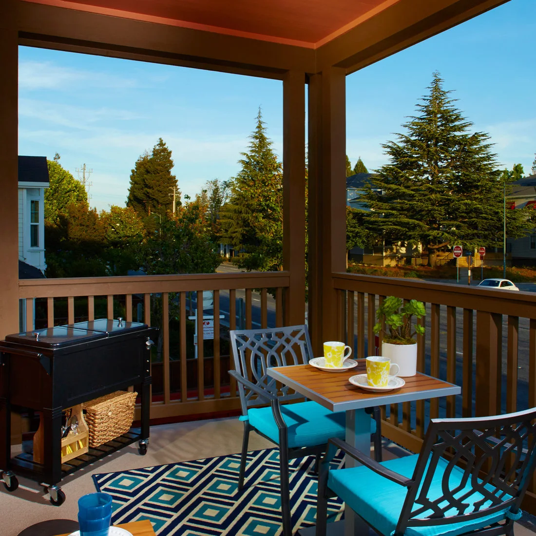 A small square bistro table and two blue-cushioned chairs sit on a covered balcony beside a serving cart