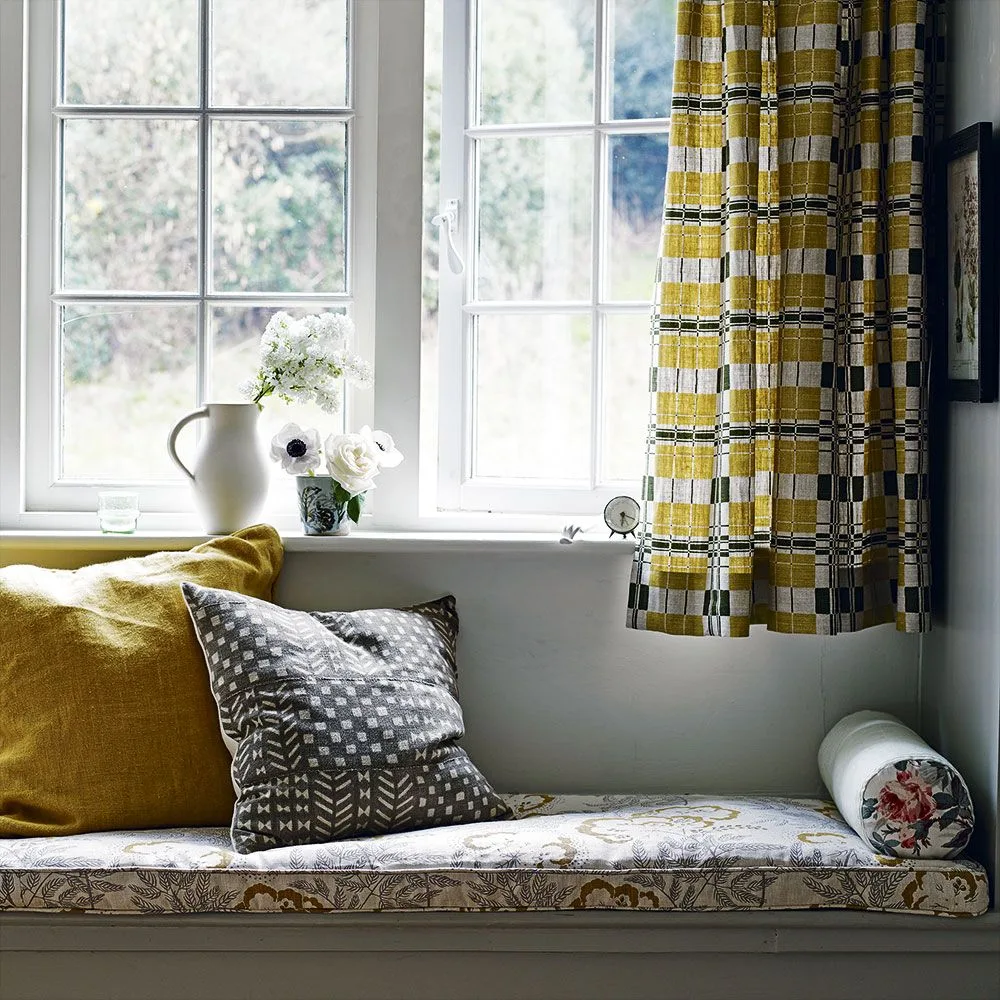 Window seat with a pale floral seat cushion, mustard and gray patterned pillows, checked curtains and flowers on the sill.