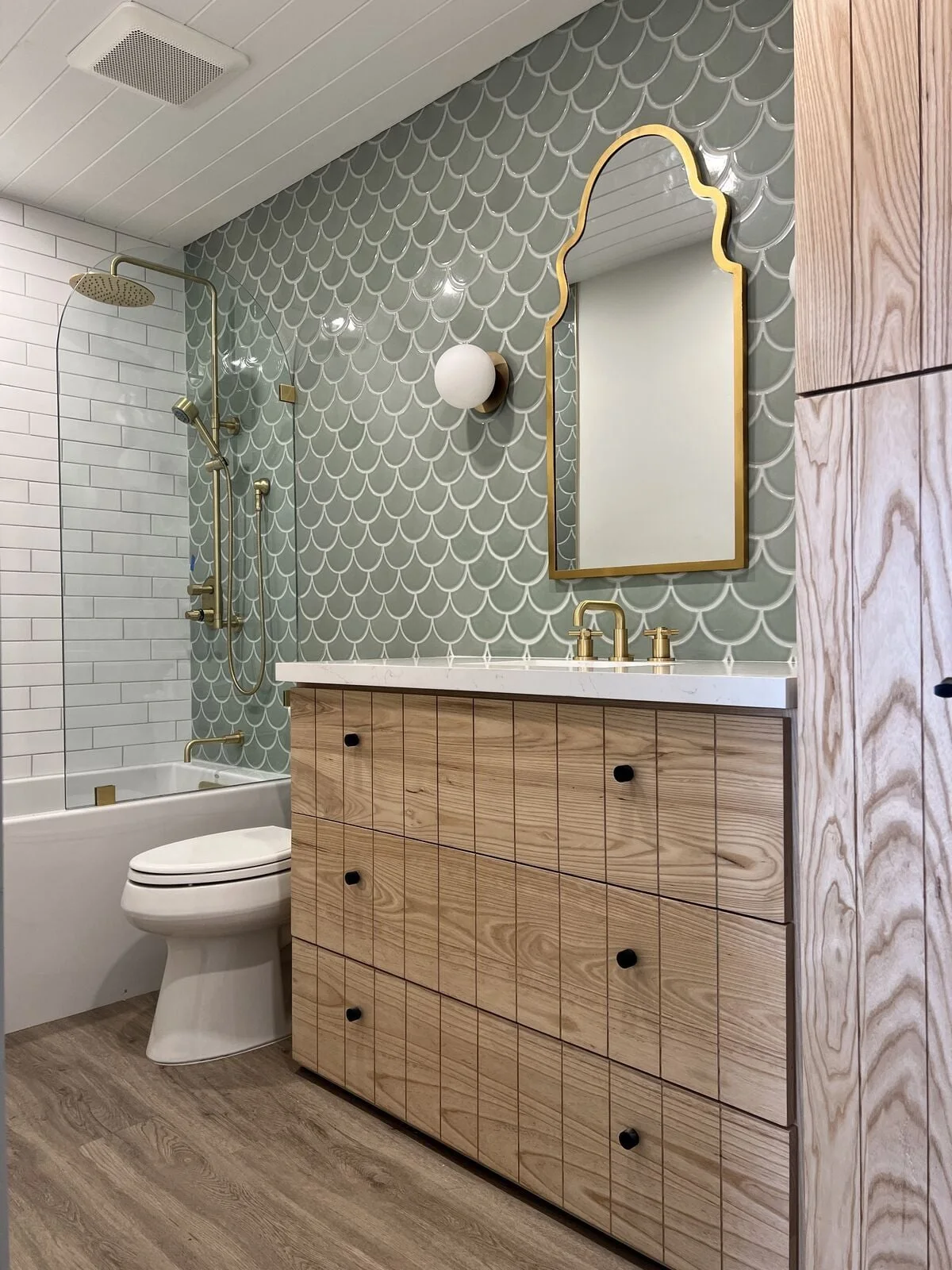 Glossy scalloped sage tiles cover a vanity wall beside white shower tile, with a shaped gold mirror and a wood drawer cabinet.