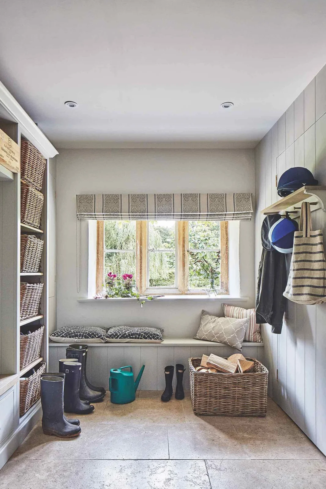 A pale entry bench below a window stands beside tall basket shelving and a coat rail against pale board walls, with boots and a large basket on the floor