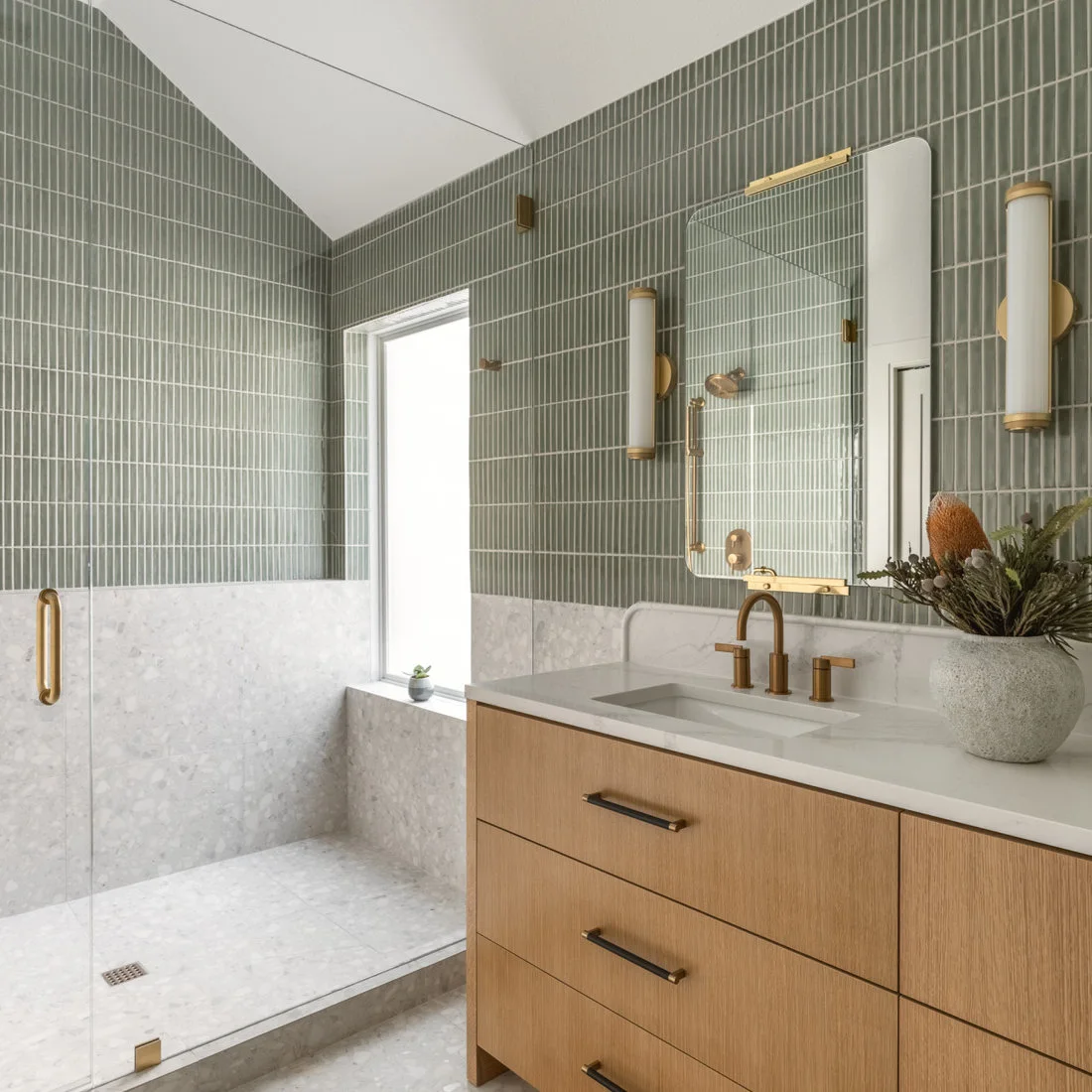 Green vertical wall tiles meet pale terrazzo beside a timber vanity and a glass shower enclosure.