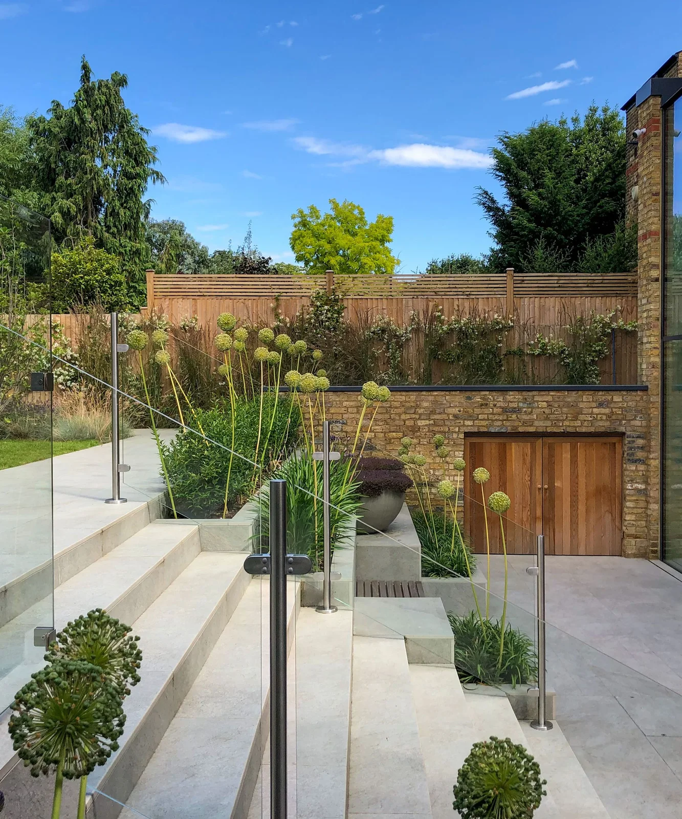 Timber storage doors sit beneath a raised brick garden area beside pale steps and planted beds